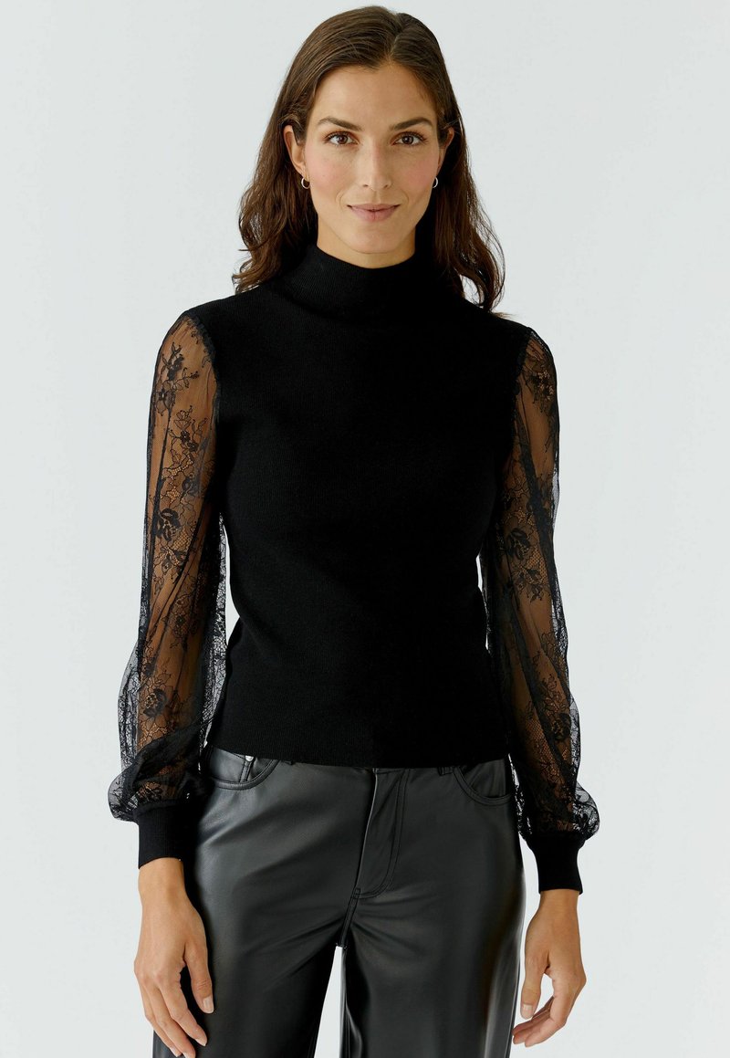 Schwarzer Pullover mit hohem Kragen, der lange, durchsichtige schwarze Spitzeärmel aufweist. Der Körper ist gestrickt, während die Ärmel florale Spitzen-Verzierungen haben.