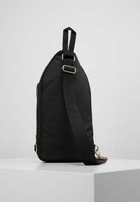 Borsa a spalla nera in tela con forma trapezoidale, dotata di una tracolla larga, accessori in oro e un dettaglio in superficie testurizzata.