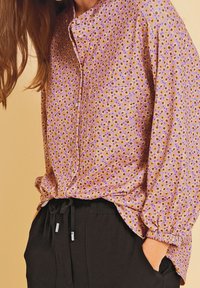 Lange mouw, lavendelkleurige blouse met klein bloemenpatroon, knoopsluiting aan de voorkant en ronde kraag. Gecombineerd met zwarte broek met elastische tailleband.