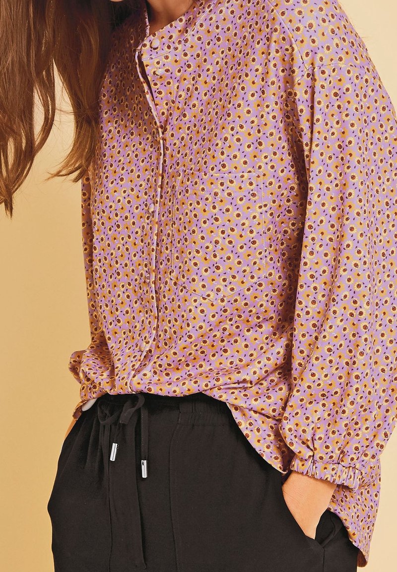 Lange mouw, lavendelkleurige blouse met klein bloemenpatroon, knoopsluiting aan de voorkant en ronde kraag. Gecombineerd met zwarte broek met elastische tailleband.