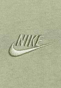 Zelená bavlněná látka je zdobena vyšívaným logem Nike ve stejném odstínu, což zdůrazňuje texturu a jemné designové detaily.