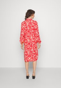 Vestido com padrão floral vermelho, de mangas longas, com comprimento até ao meio da perna, cintura ajustada e um corte descontraído. Tecido texturizado; a vista traseira mostra o fecho por botão.