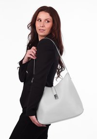 Šedá kožená taška na rameno s hladkým povrchem, zaobleným tvarem a stříbrným akcentem na popruhu. Taška je držena přes rameno.