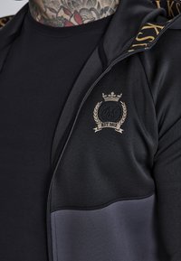 Černá sportovní bunda se zipem vpředu, zlatými detaily loga a hladkou texturou. Obsahuje šedou dolní část a kontrastní podšívku kapuce.