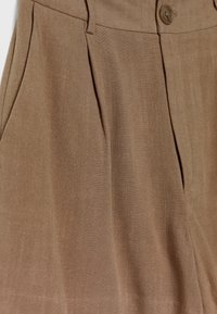 Pantalon marron sur mesure en tissu texturé, avec fermeture à boutons, plis et poches de chaque côté.