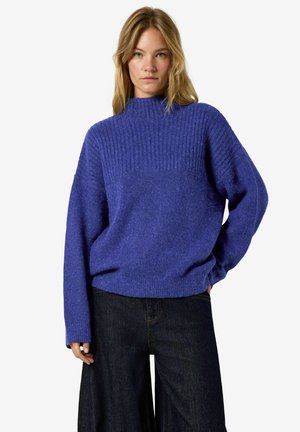 Femme portant un pull en maille côtelée violet ample à manches longues et un jean large foncé, debout avec la main dans la poche.