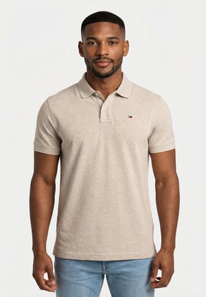 Man met kort haar en baard, gekleed in een beige polo met korte mouwen en lichtblauwe spijkerbroek, staand tegen een effen witte achtergrond.
