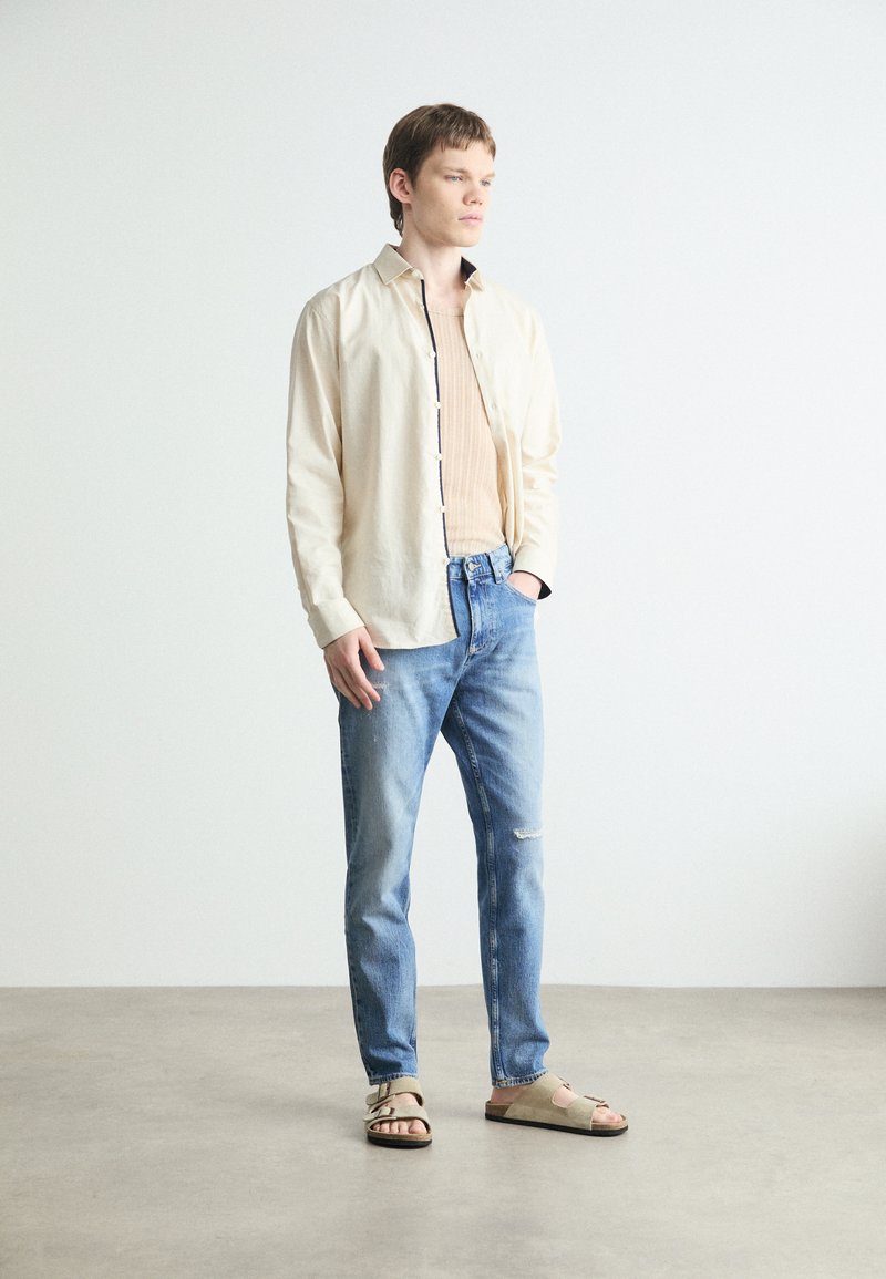 Hellbeige Hemd mit Knöpfen, gerippte hellbraune Tanktop, ausgewaschene blaue Jeans und graue Sandalen mit Schnalle. Sanfte Texturen und lässige Passform.