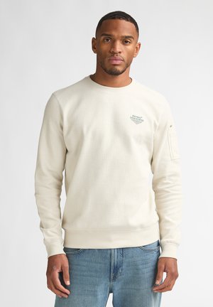 Homme portant un sweat-shirt crème à col rond avec un petit logo "Petrol Industries" sur la poitrine et un jean bleu, debout devant un fond blanc uni.