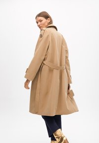Beige trenchcoat med bälte i midjan, stor krage och knappdetaljer. Har lång ärm och en öppning i ryggen för ökad rörelsefrihet.