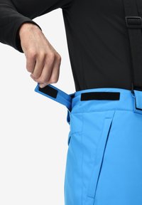 Een hand die een zwarte klittenbandsluiting aanpast op een felblauwe waterdichte broek, gedragen met een zwart longsleeve shirt.