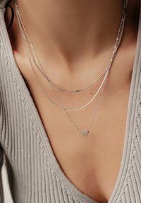 Drei geschichtete Silberketten; die niedrigste zeigt einen kleinen Herzanhänger. Im Hintergrund ist ein strukturiertes graues Kleidungsstück sichtbar.