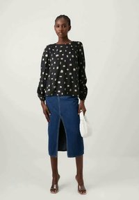 Ženska nosi črno dolgosleeve vzorčasto bluzo, modro krilo iz denima do kolen s sprednjim razporkom, rjave sandale na peto, drži belo torbico.