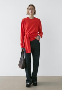 Rode trui met een knoopdetail in de taille, gecombineerd met donkere, wijde broekspijpen. Bruine leren tas en zwarte platte schoenen. Eenvoudig, modern ontwerp.