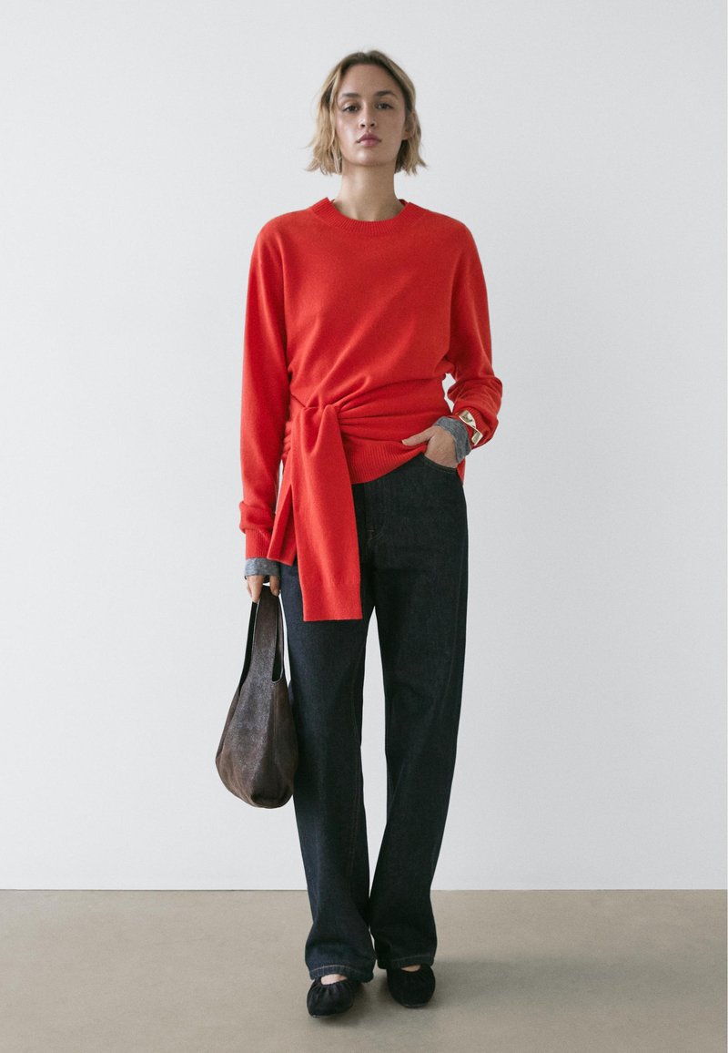 Rode trui met een knoopdetail in de taille, gecombineerd met donkere, wijde broekspijpen. Bruine leren tas en zwarte platte schoenen. Eenvoudig, modern ontwerp.