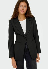 Must mängitud must bleiser ühe nupuga kinnitusega, nurgeliste kraedega ja kitsas lõige. Omab küljetaskuid ja struktureeritud õlgu.