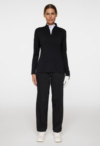 Vrouw staat rechtop in een zwart opstaande kraag lange mouw top, zwarte broek, witte handschoen om haar linkerhand en witte sneakers tegen een effen achtergrond.