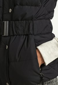 Czarna pikowana kurtka z szerokim elastycznym paskiem i srebrną klamrą, ręka w bocznej kieszeni, widoczny kremowy ściągacz na rękawie.