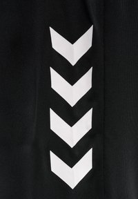 Sorte træningsbukser med en række hvide chevron-mønstre langs siden, lavet af et glat, strækbart stof.