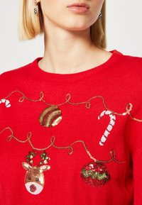 Rode gebreide trui met paillettenversieringen van een rendier, een zuurstok en kerstballen op een ontwerp van gouden draad.