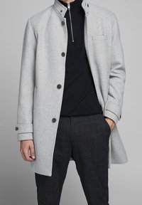 Abrigo de lana gris claro con cuello alto, cierres de botón, un bolsillo en el pecho y una textura suave, combinado con pantalones oscuros de cuadros.