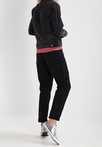 Giacca di jeans nera con texture sbiadita, abbinata a una camicia rossa e pantaloni neri. Indossata con scarpe da ginnastica silver, mostra un outfit casual visto da dietro.