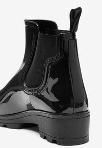 Botte Chelsea en caoutchouc noir brillant avec panneau latéral élastique, languette arrière et semelle extérieure texturée noire. Bout arrondi et finition lisse.