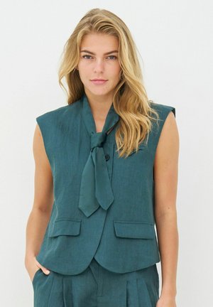 Femme blonde portant un gilet sans manches sarcelle avec une cravate assortie et un pantalon, se tenant avec une main dans la poche devant un fond uni.