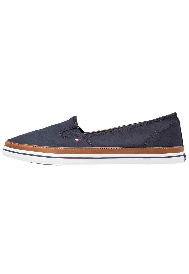 Navy canvas slip-on kengät ruskeilla nahkaakatsomuksilla, valkoisella kumipohjalla ja ommelluilla yksityiskohdilla. Minimalistinen muotoilu elastisilla sivupaneeleilla.