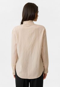 Lichtbeige langemouwen shirt met een subtiele gestructureerde print, met knoopmanchetten en een gebogen zoom. De achterkant laat een ontspannen pasvorm en een kraag zien.