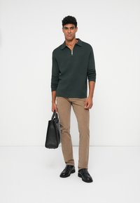 Grønn langermet polo-skjorte med glidelås i kragen, sammen med brune bukser og svarte loafers. Modellen holder en svart skinn totebag.