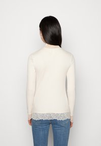 Langermet kremfarget ribbet topp med blondekant ved halsen og nederkant, med en tettsittende design og myk tekstur.