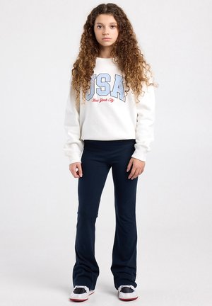 Meisje met lang krullend haar, gekleed in een wit USA-sweatshirt, marineblauwe wijde broek en zwart-witte sneakers, staand tegen een effen achtergrond.
