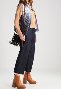 Vadderad silverväst, beige stickad tröja, mörk denim bredbenta byxor, bruna ankelstövlar med kraftiga klackar och en fransad svart handväska.