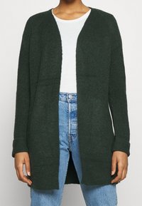 Žena v tmavě zeleném kardiganu s otevřeným předním dílem, pod kterým má bílé tričko, a v modrých kalhotách s vysokým pasem, stojící před jednobarevným pozadím.