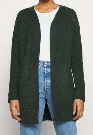 Woman wearing dark green open-front cardigan over white top and blue high-waisted jeans, standing against a plain background.