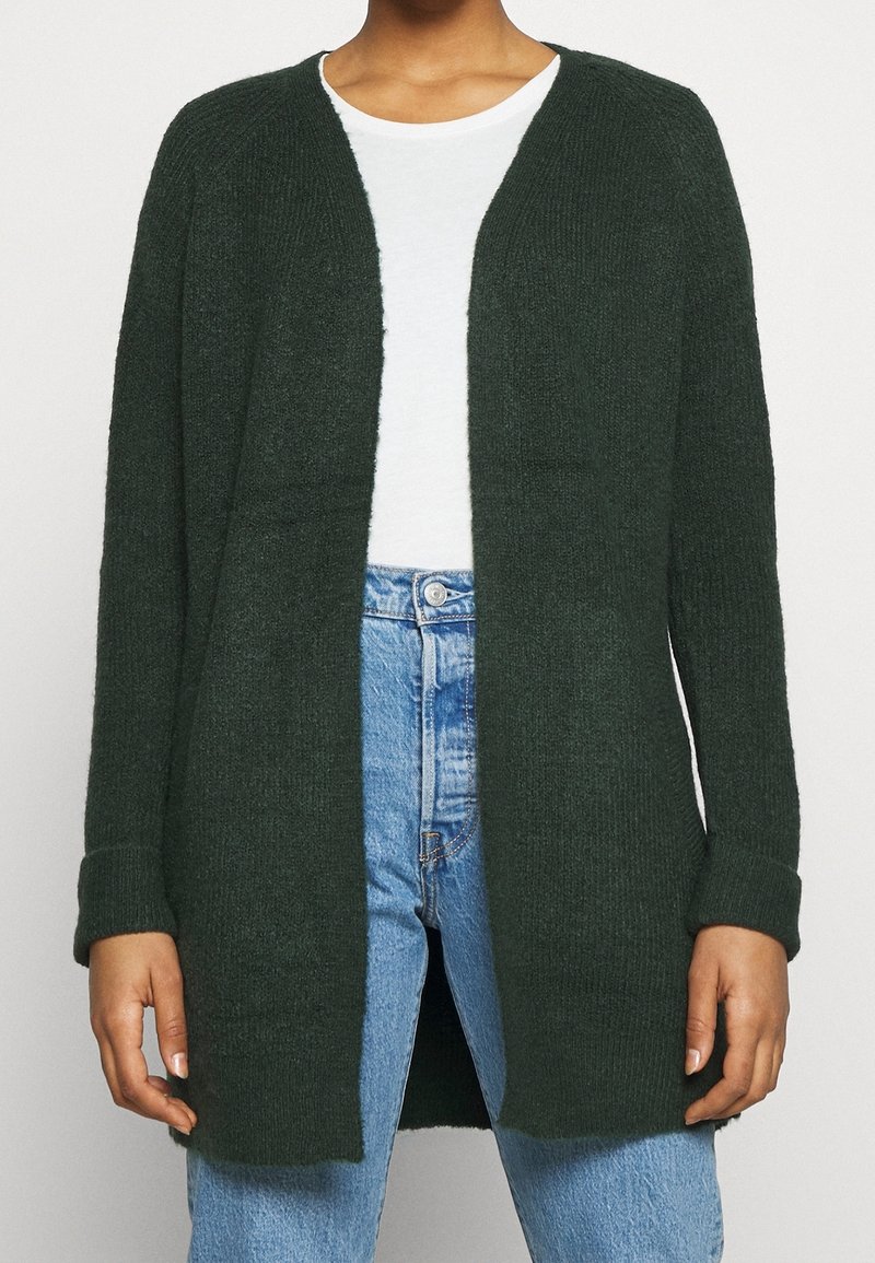 Žena v tmavě zeleném kardiganu s otevřeným předním dílem, pod kterým má bílé tričko, a v modrých kalhotách s vysokým pasem, stojící před jednobarevným pozadím.