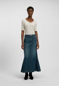 Vaaleanbeige lyhythihainen toppi, jossa nappitolppa kaula-aukossa, yhdistettynä pitkään, liehuvaan tummansiniseen denimhameeseen. Mallilla on jalassaan mustat nilkkurit.