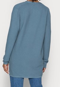 Dlouhý rukáv tunika ve světle modré barvě s žebrovanou texturou, kulatým výstřihem a délkou po boky. Pohodlný a neformální střih.