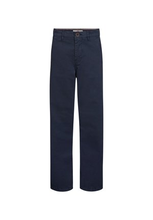 Jongen met krullend haar staat en draagt een lichtgrijs shirt met lange mouwen, donkerblauwe broek en witte sneakers tegen een eenvoudige lichte achtergrond.