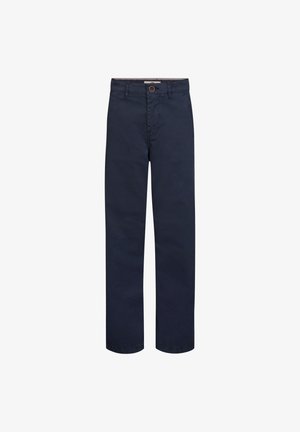 Jongen met krullend haar staat en draagt een lichtgrijs shirt met lange mouwen, donkerblauwe broek en witte sneakers tegen een eenvoudige lichte achtergrond.