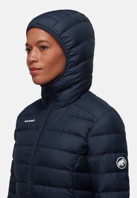 Tamno plava izolirana puffer jakna s uskim kapuljačom, zatvaranjem na zip i Mammut logotipom na rukavu. Kvadratna tekstura s horizontalnim šavovima.