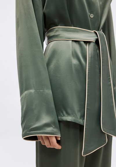 Πράσινο σατέν τοπ με μακριά μανίκια, φαρδιά μανσέτα και μπεζ περιγράμματα, με μια ταιριαστή ζώνη δεμένη στη μέση πάνω από πράσινο παντελόνι.