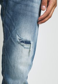 Vaaleansiniset kulutetut farkut, joissa on iso repeämä oikeassa reidessä, kuluneet reunat ja haalistunut pesu.