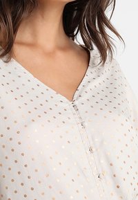 Blusa grigia chiara con scollo a V e motivo a pois dorati. Presenta bottoni sul davanti e una morbida consistenza del tessuto che scivola.