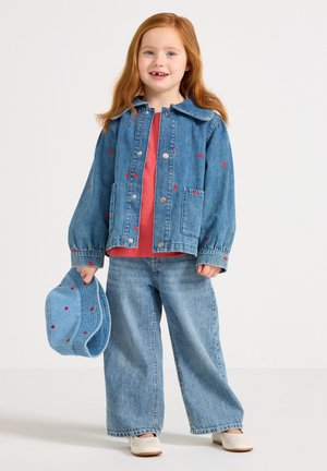 Nuori punatukkainen tyttö, jolla on päällään farkkutakki ja housut, joissa on punaisia sydänkuvioita, pitelee vastaavaa lakkia, hymyilee, seisoo valkoista taustaa vasten.