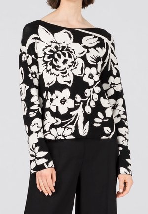 Zwarte trui met lange mouwen en groot wit bloemmotief, gedragen met zwarte broek, getoond van schouders tot heupen.