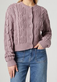 Lila kabelstickad kofta med rund hals, sex matchande knappar och ribbad nederkant och ärmslut, kombinerad med ljusblå jeans.