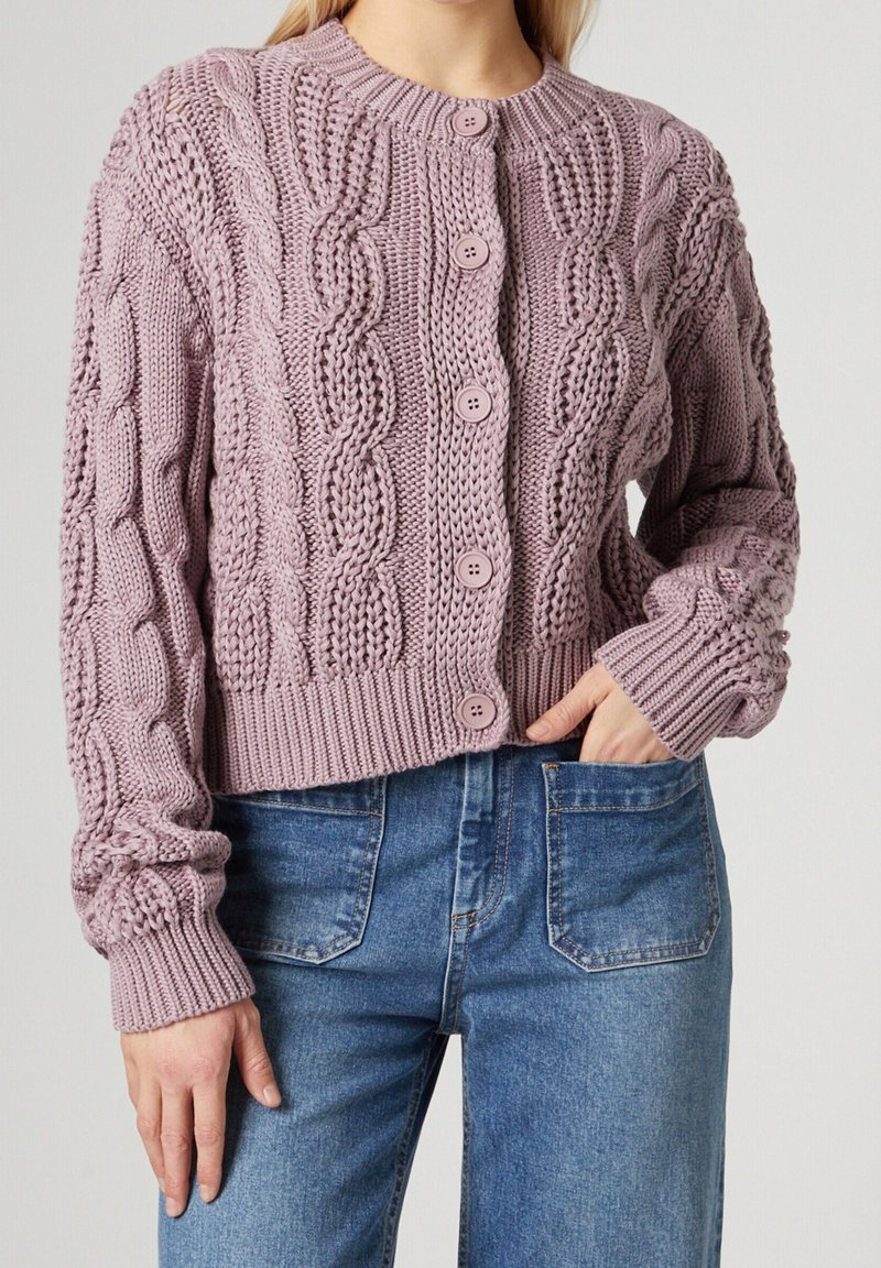 Lila kabelstickad kofta med rund hals, sex matchande knappar och ribbad nederkant och ärmslut, kombinerad med ljusblå jeans.