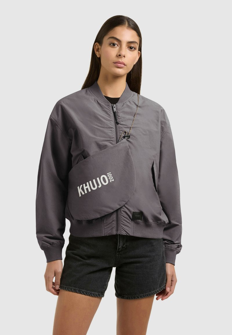 Jeune femme portant une veste bomber grise zippée, un short en jean noir et un sac bandoulière gris avec le texte "KHUJO 2001".